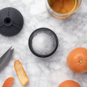 Round ice cube mold with ice surrounded by oranges and orange peels and a glass