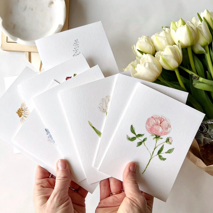 Set of watercolor floral cards held by hands with a bouquet of white tulips in the background.