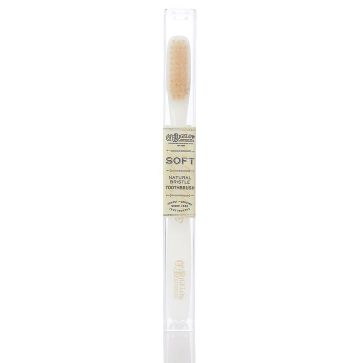 A white toothpaste with pale bristles by c o bigelow in a container with a label reading "soft natural bristle toothbrush"
