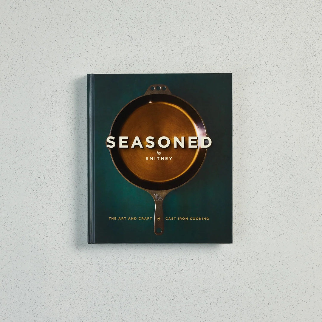 The book 'Seasoned by Smithey: The Art and Craft of Cast Iron Cooking' is displayed flat against a light gray textured background. The cover features a dark teal-green background with a central illustration of a cast iron skillet showing a golden-brown interior, with the title in white text.