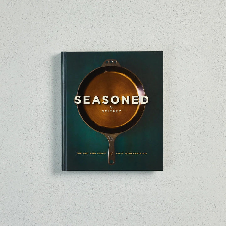 The book 'Seasoned by Smithey: The Art and Craft of Cast Iron Cooking' is displayed flat against a light gray textured background. The cover features a dark teal-green background with a central illustration of a cast iron skillet showing a golden-brown interior, with the title in white text.
