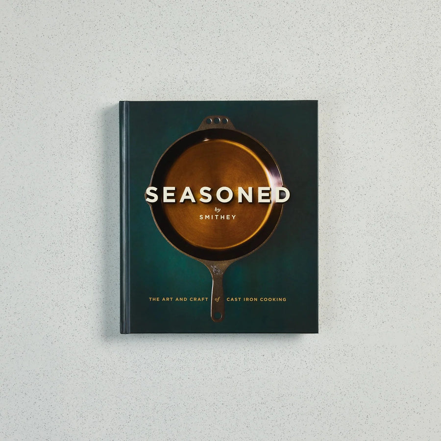The book 'Seasoned by Smithey: The Art and Craft of Cast Iron Cooking' is displayed flat against a light gray textured background. The cover features a dark teal-green background with a central illustration of a cast iron skillet showing a golden-brown interior, with the title in white text.