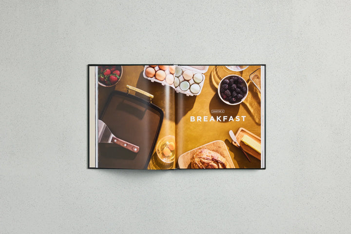 An open cookbook or recipe book displayed against a light gray textured wall. The spread shows a breakfast food scene with eggs, berries, bread, and a baking pan, with the word 'BREAKFAST' printed across the pages.