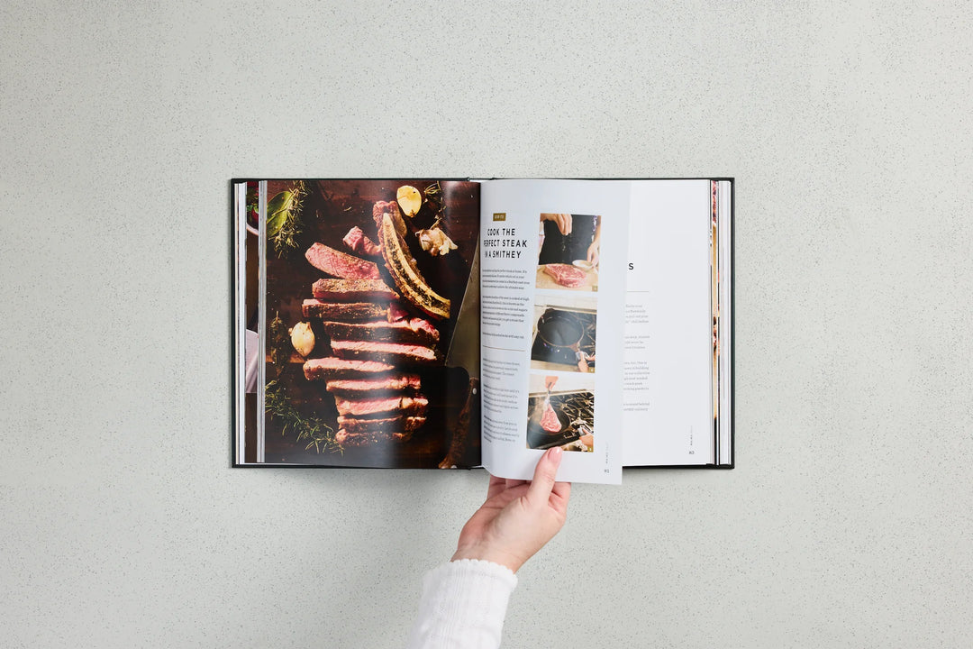 An open cookbook or recipe book is laid flat against a light gray textured background, with a hand holding the right page. The left page shows a close-up photo of stacked ribs with lemon garnish, and the right page contains text and small food photography panels.