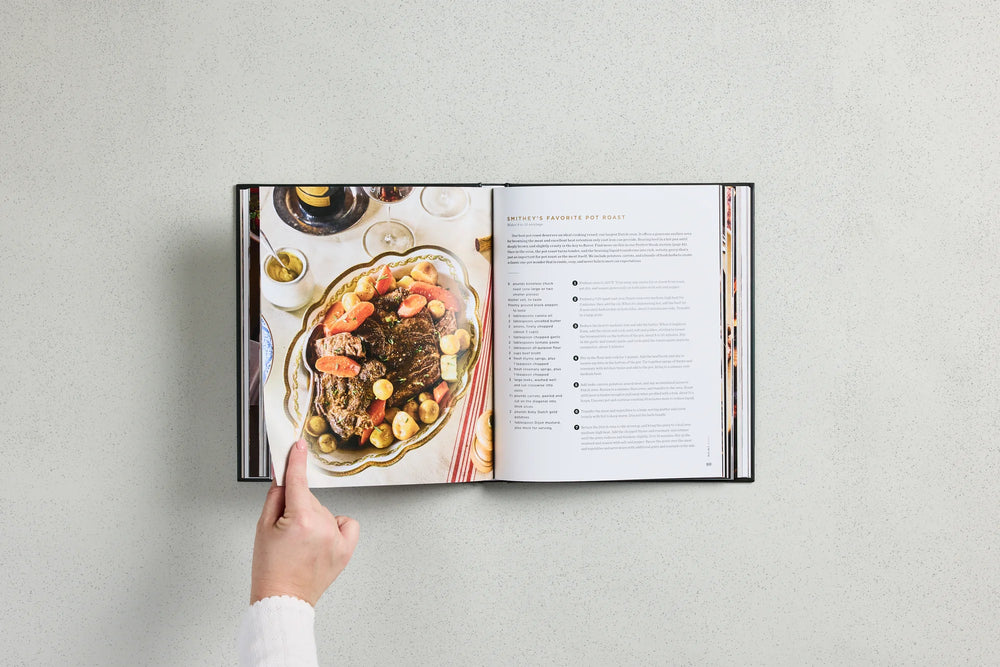 An open hardcover cookbook lies on a light gray surface, showing a plated steak dish on the left page and recipe text on the right. A person's hand points toward the cookbook.