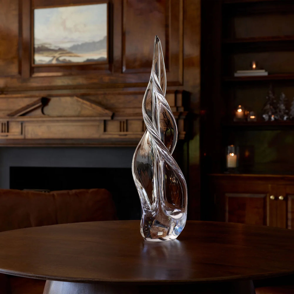 A clear glass christmas tree sculpture with a twisted, flame-like form stands on a dark wooden table in a warm, dimly lit room with wood paneling and a fireplace in the background.