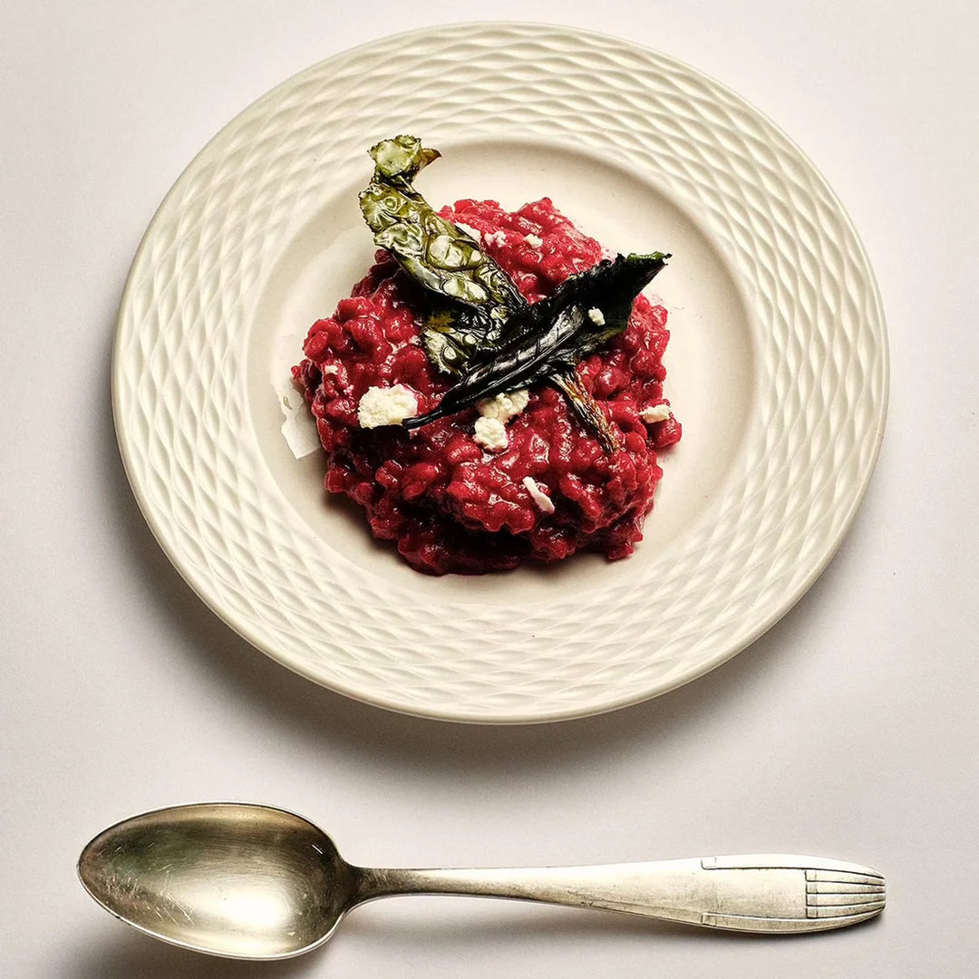 A plated food dish showing a mound of dark red risotto with a leaf garnish and small white crumbles, served on a textured off-white plate with a silver spoon below on a light gray surface.