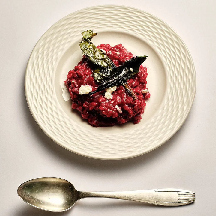 A plated food dish showing a mound of dark red risotto with a leaf garnish and small white crumbles, served on a textured off-white plate with a silver spoon below on a light gray surface.