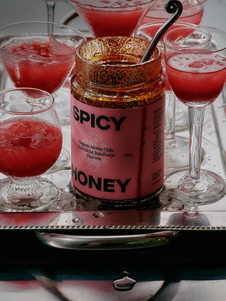 A jar of 'Spicy Honey' chili-infused honey sits on a silver tray surrounded by glasses of a pink-red chili cocktail.