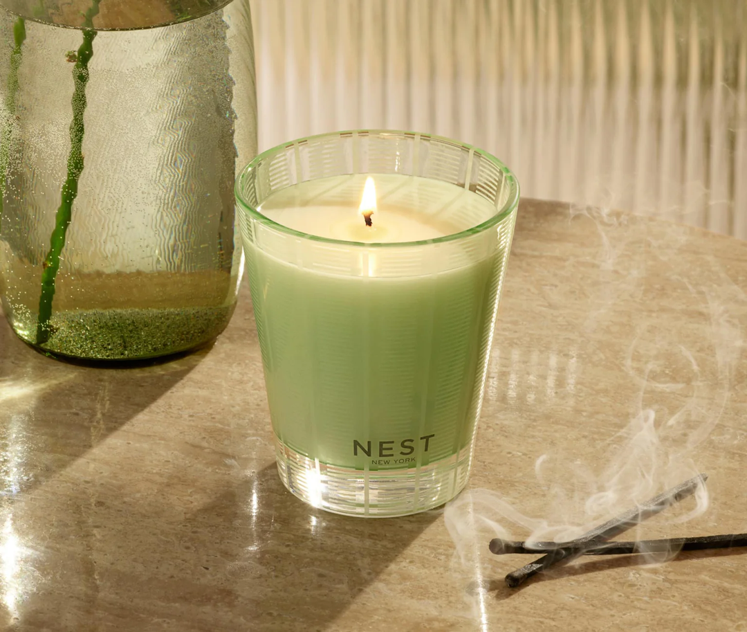 GreenNest Cucumber and White sage candle on a pale marble table nest to a few smoking matches and a green glass vase with flower stems