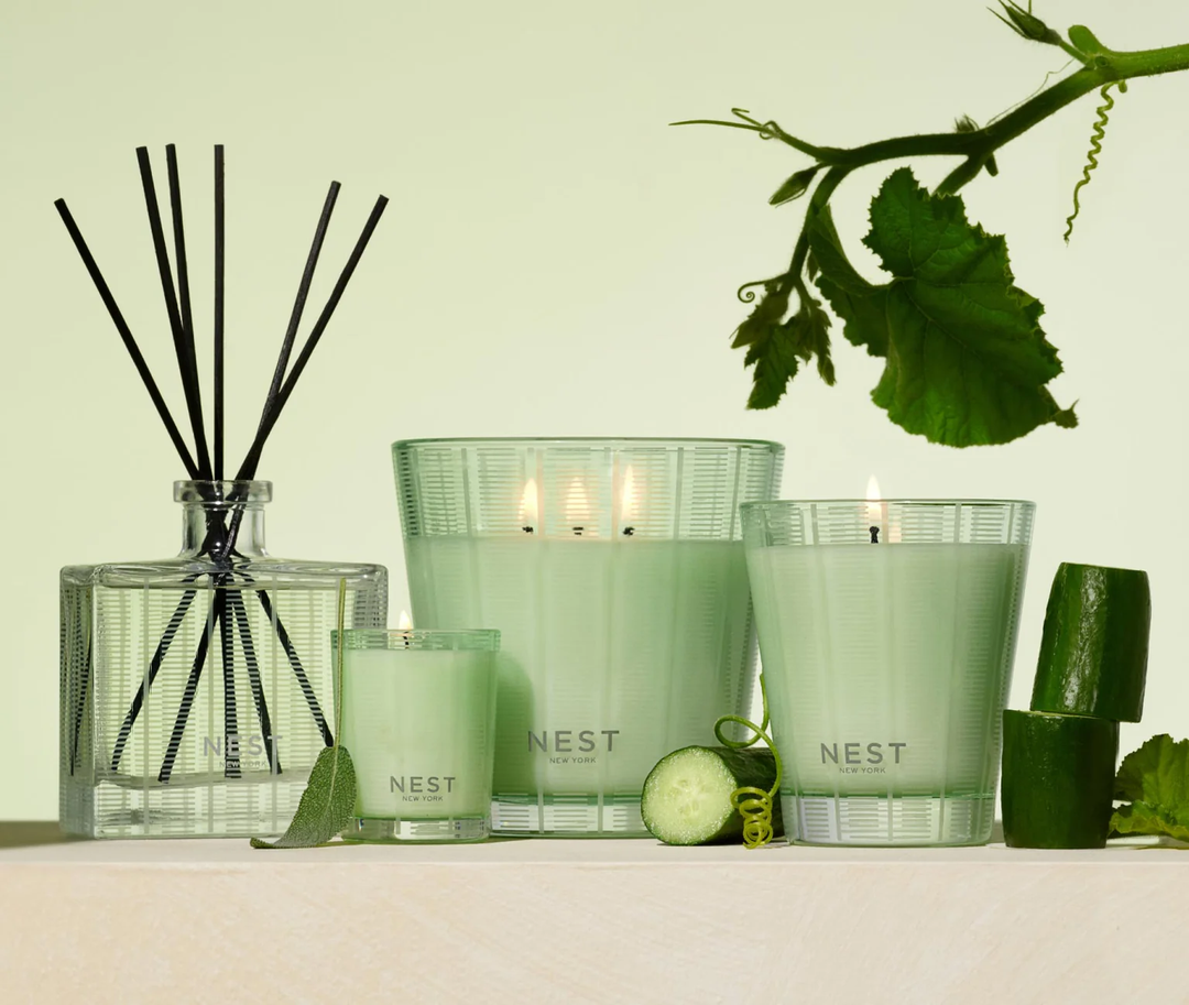 Green nest cucumber and white sage votice, classic, and three wick candles with a reed diffuser sitting ith decorative cucumbers and vines