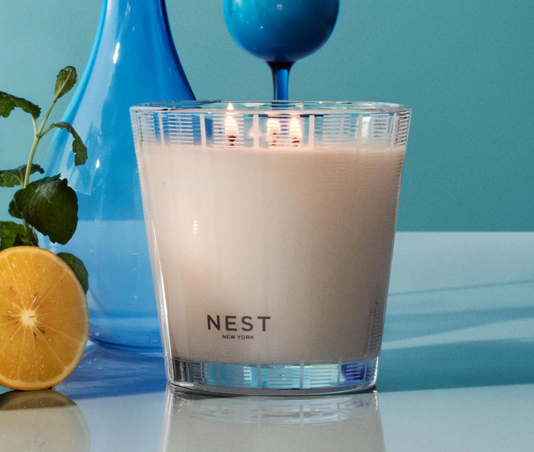 nest amalfi lemon and mint three wick candle on a reflective surface with lemons and mint and blue vases with a blue background