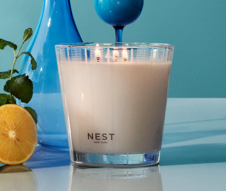 nest amalfi lemon and mint three wick candle on a reflective surface with lemons and mint and blue vases with a blue background