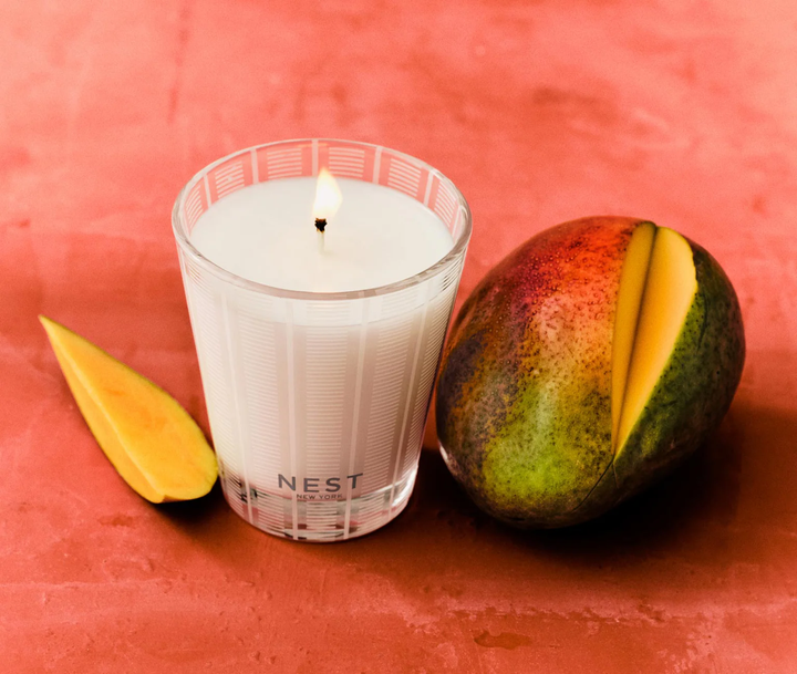 A nest Something Tropical Candle with a sliced mango on an orange red surface