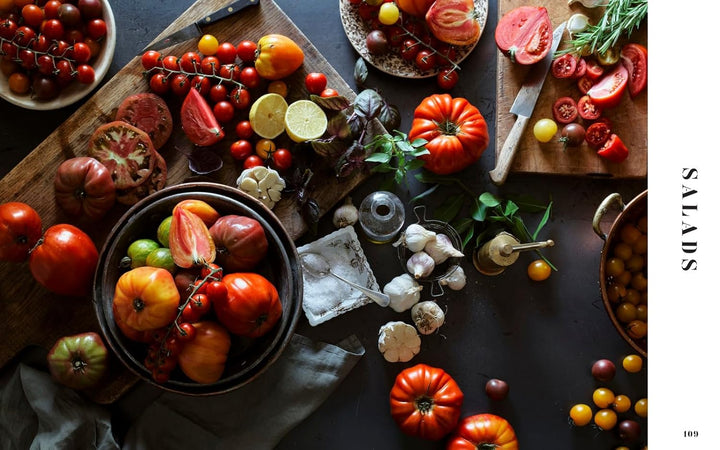 A top view of a spread of cooking ingredients featuring tomatos, garlic, salt, paper grinders, and knives