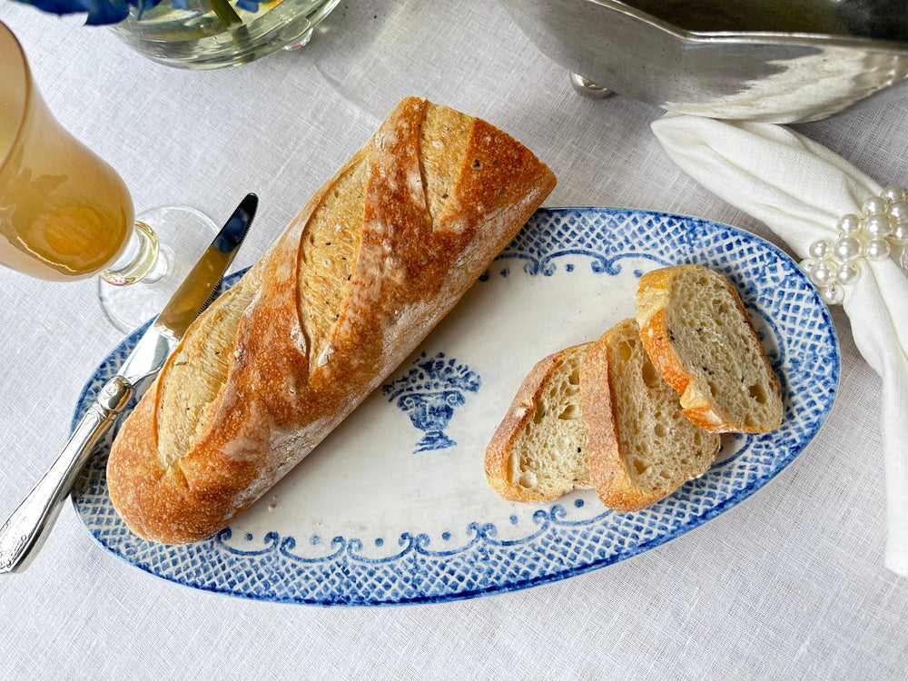 A rustic loaf of bread with several slices on a blue and white patterned oval platter, set on a white tablecloth with a glass, knife, and napkin nearby.