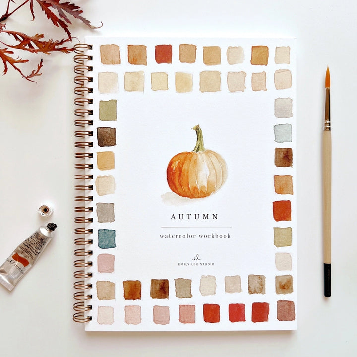 A watercolor book with a pumpkin illustration surrounded by paint swatches on the cover sitting beside a paintbrush