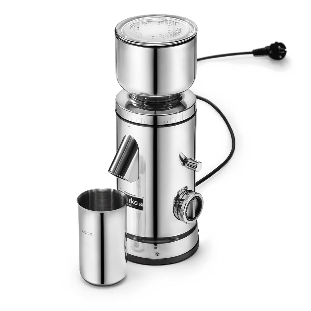 A stainless steel electric grinder with a cylindrical body, a top hopper, a side chute, a small metal cup, and a power cord, shown on a clean white background.
