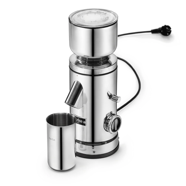 A stainless steel electric grinder with a cylindrical body, a top hopper, a side chute, a small metal cup, and a power cord, shown on a clean white background.