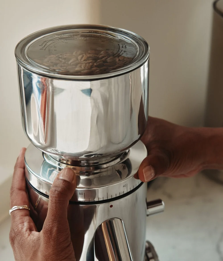 A close-up of a stainless steel coffee grinder with a clear bean hopper filled with coffee beans, held by two hands against a soft beige background.