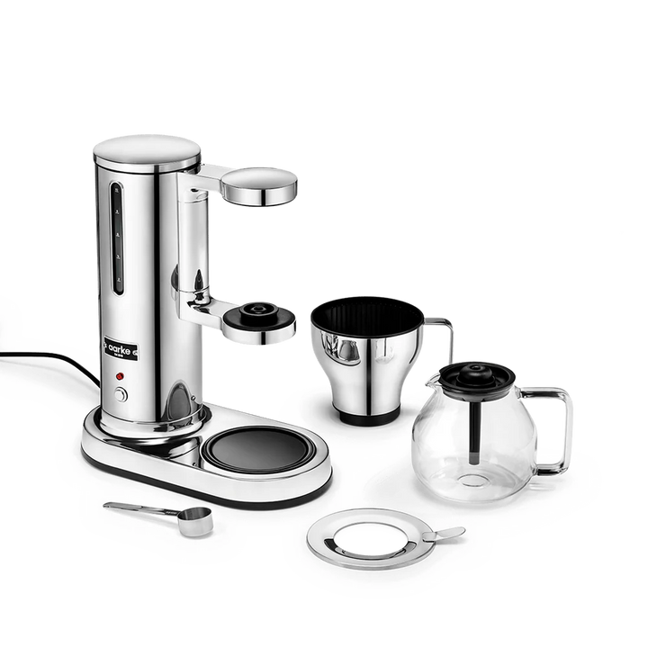 A stainless steel espresso machine set with a glass carafe, metal filter basket, measuring scoop, and lid, arranged on a clean white background.