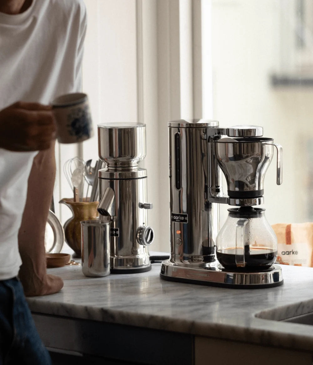 Person holding a coffee cup next to a coffee maker on a kitchen counter.