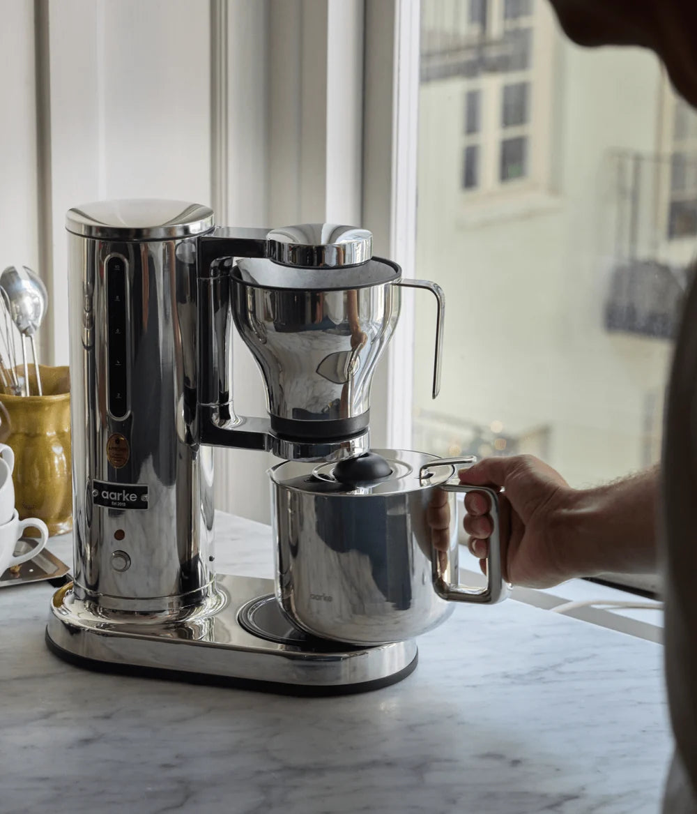 Stainless steel coffee maker on a marble countertop with a person's hand holding the carafe.