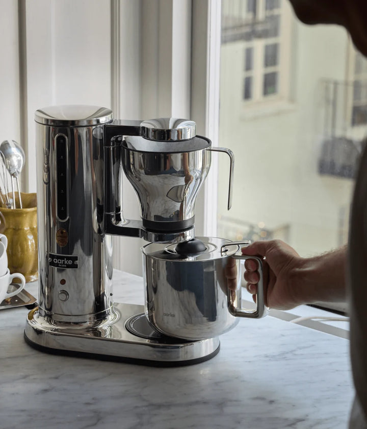 Stainless steel coffee maker on a marble countertop with a person's hand holding the carafe.