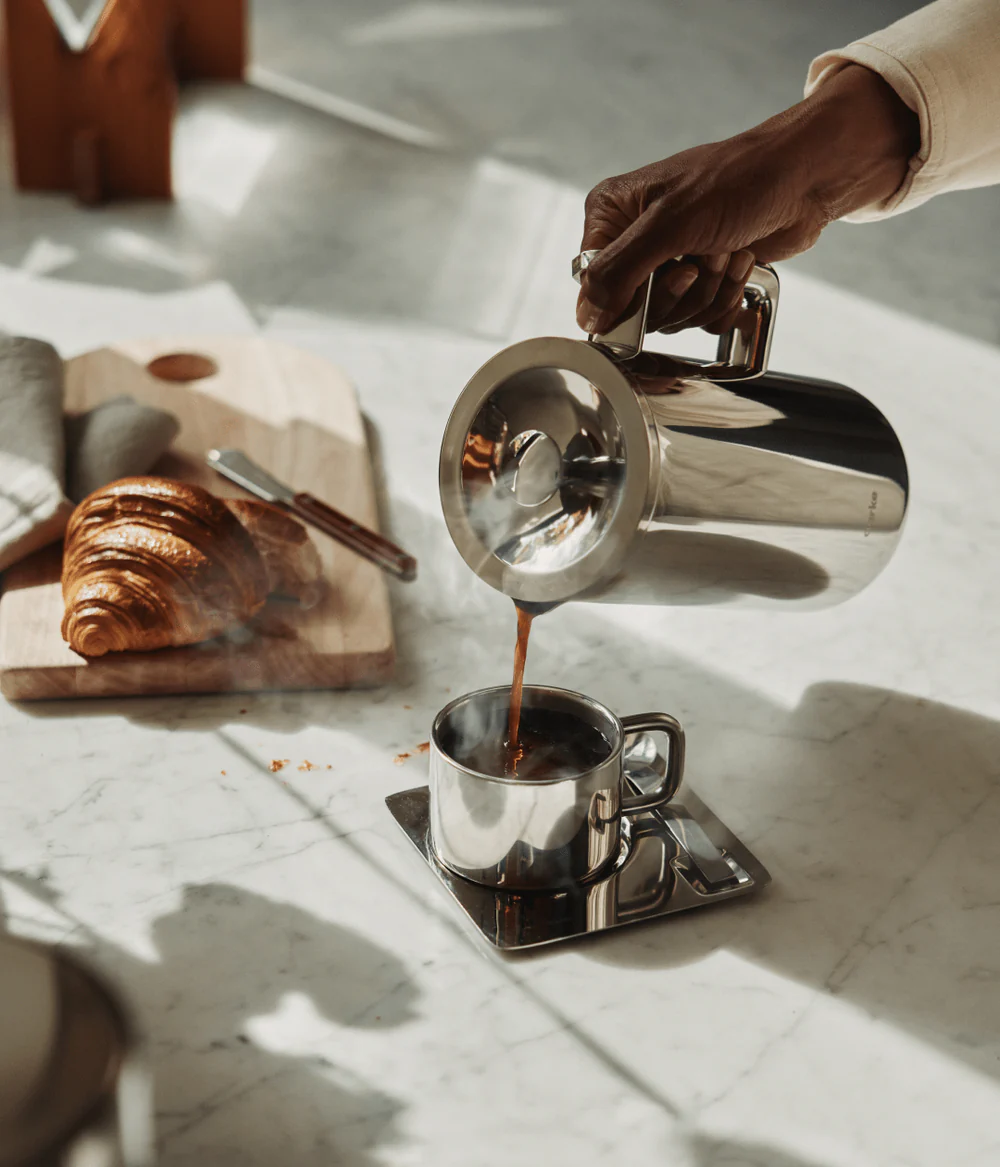 Person pouring coffee from a silver kettle into a cup on a marble surface with a croissant and wooden cutting board.