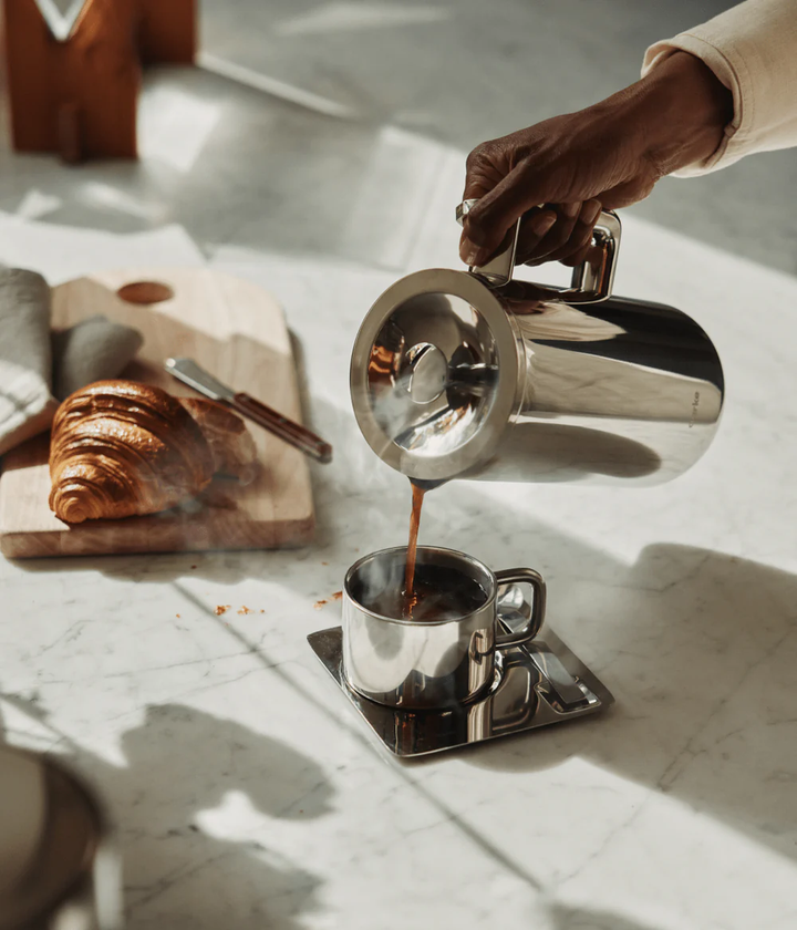 Person pouring coffee from a silver kettle into a cup on a marble surface with a croissant and wooden cutting board.