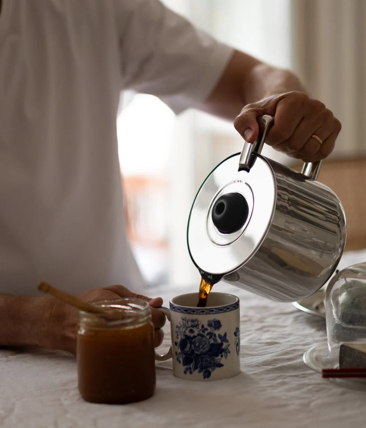 Person pouring coffee from a silver kettle into a white mug with blue floral design on a bed.