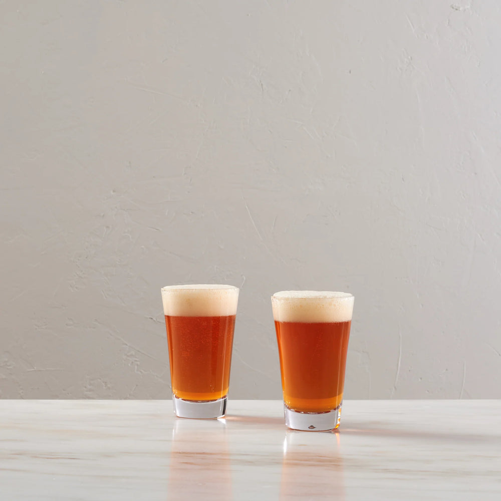 Two small clear shot glasses filled with amber beer and topped with thick white foam, standing on a light stone surface against a textured light gray wall.