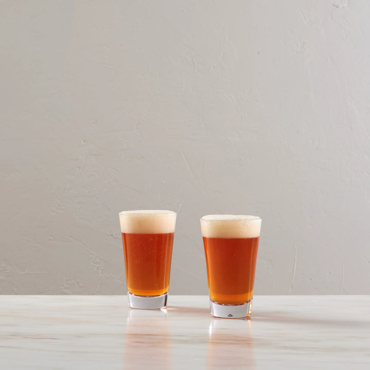 Two small clear shot glasses filled with amber beer and topped with thick white foam, standing on a light stone surface against a textured light gray wall.