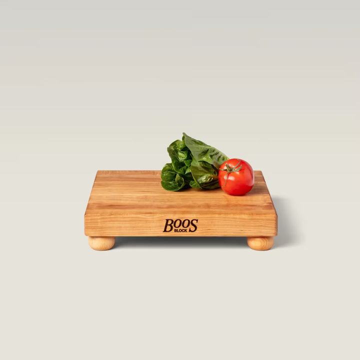 A rectangular wooden cutting board with small round feet, branded 'BOOS MADE IN USA' on the front, shown on a light gray studio background with a green leafy herb and a red tomato placed on top.