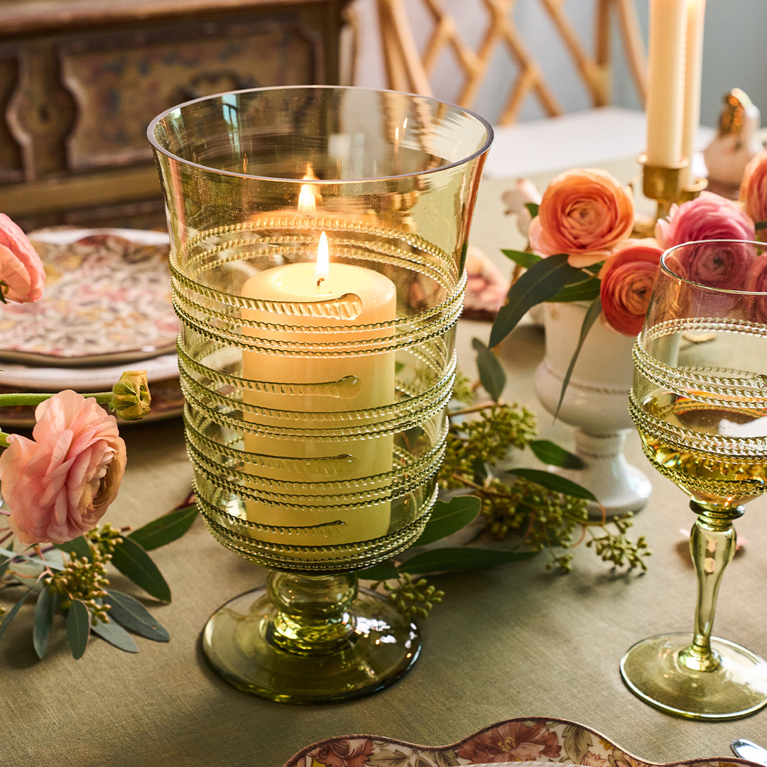 Decorative candle in a glass holder on a table with flowers and a glass of wine.