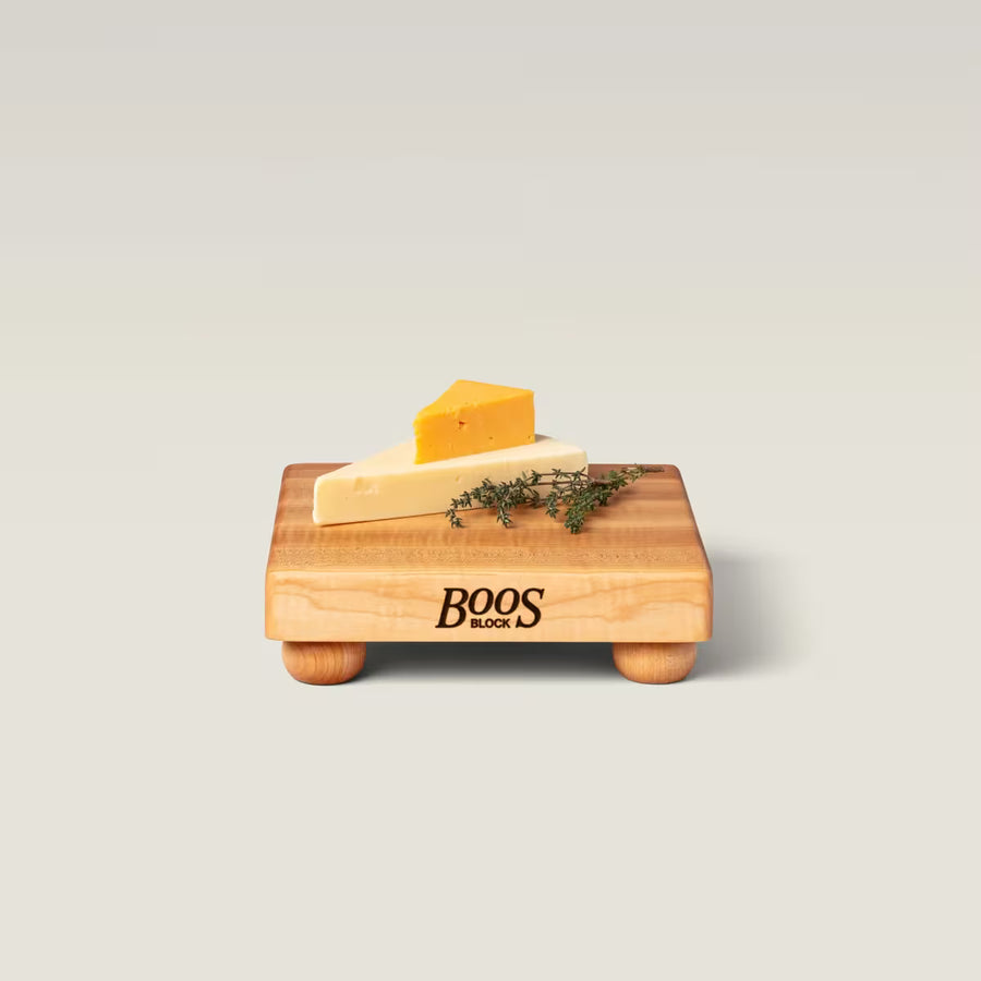 A small wooden cheese board with rounded feet on a light gray background. The board is branded 'BOOS BLOCK' and is styled with a wedge of white cheese, a smaller wedge of orange cheddar, and a sprig of fresh thyme.
