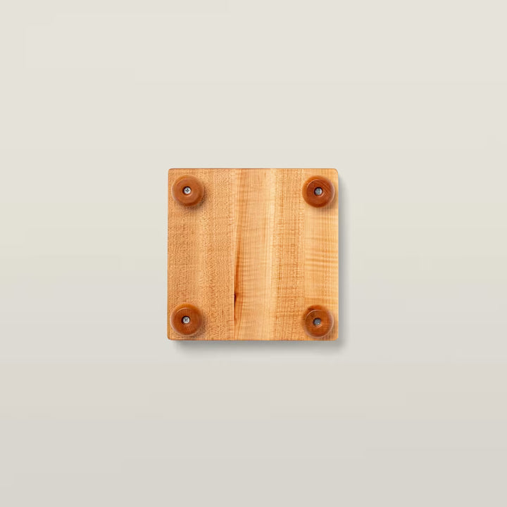 A square wooden board with a natural light brown grain and four small circular brown rubber feet, shown from a top-down view on a light beige background.