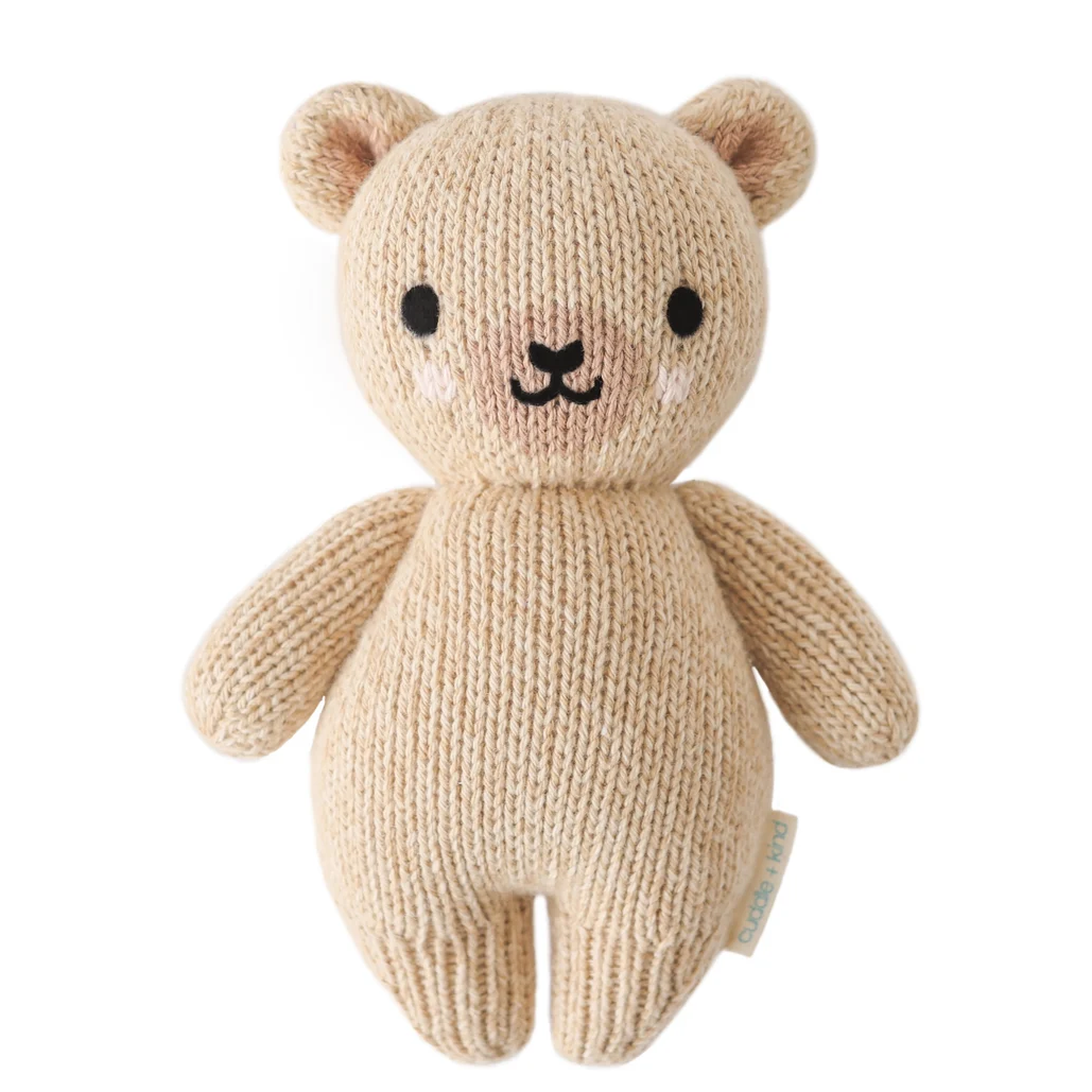 A beige knitted teddy bear plush toy with black embroidered eyes, nose, and mouth