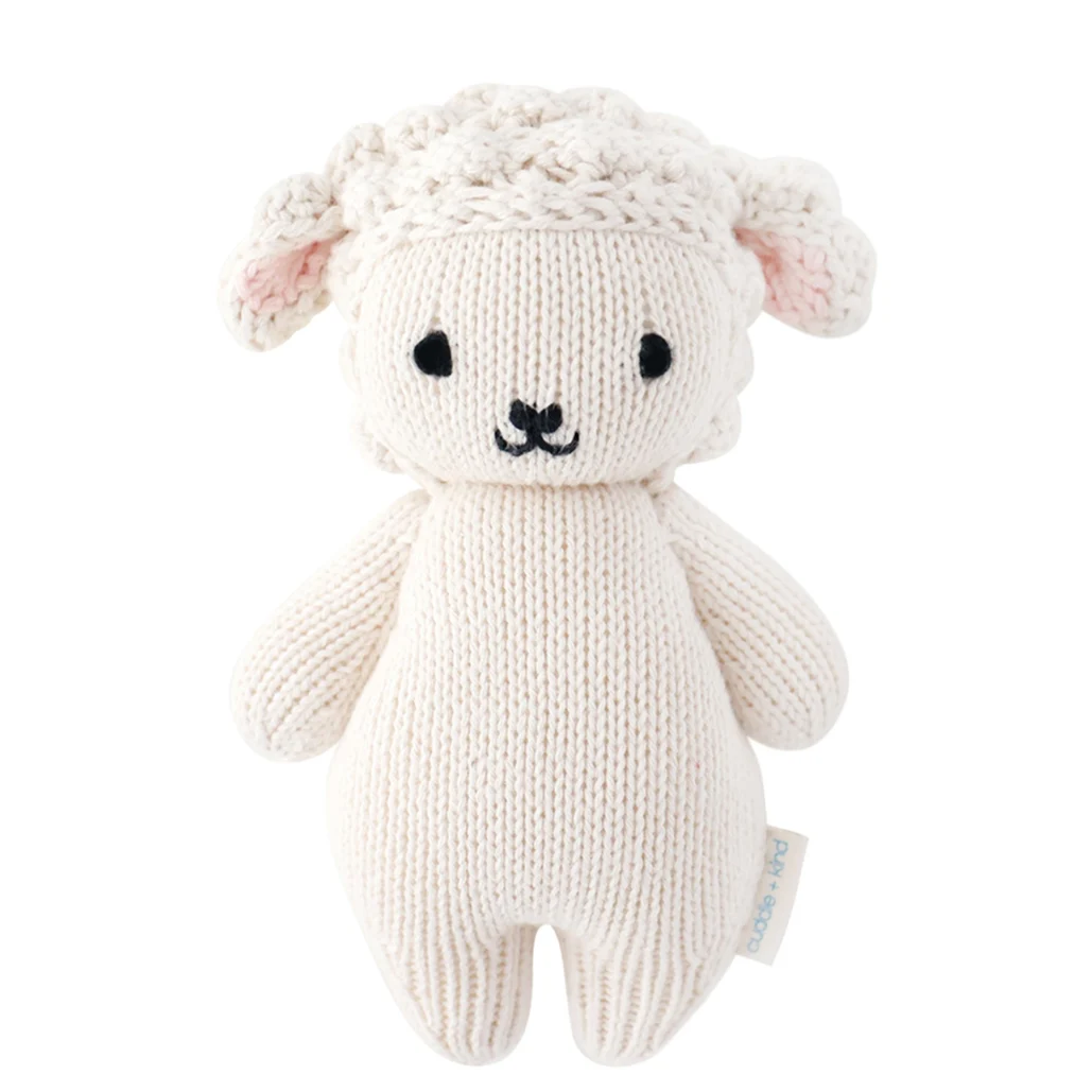 A cream-colored knitted sheep toy with black embroidered eyes and nose, pink inner ears, and a small fabric tag on the side