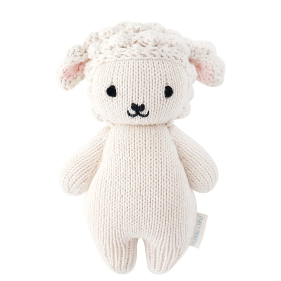 A cream-colored knitted sheep toy with black embroidered eyes and nose, pink inner ears, and a small fabric tag on the side