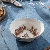 A scalloped ceramic bowl with brown botanical and house motifs, set on a blue tablecloth with dining glassware visible around it.