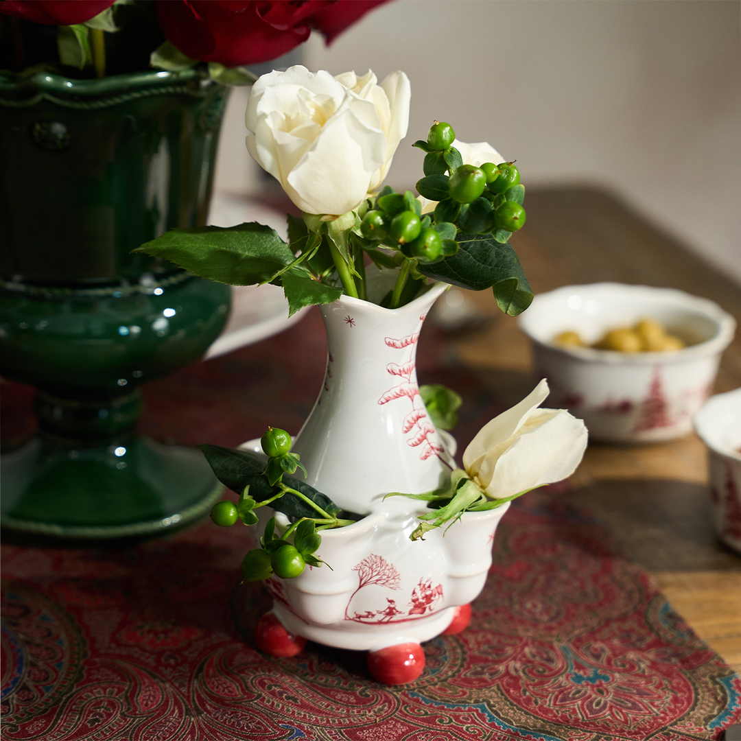 A small white ceramic vase with red floral illustrations and red feet, holding white roses and green berries, styled on a patterned red table runner with other decorative vases in the background.