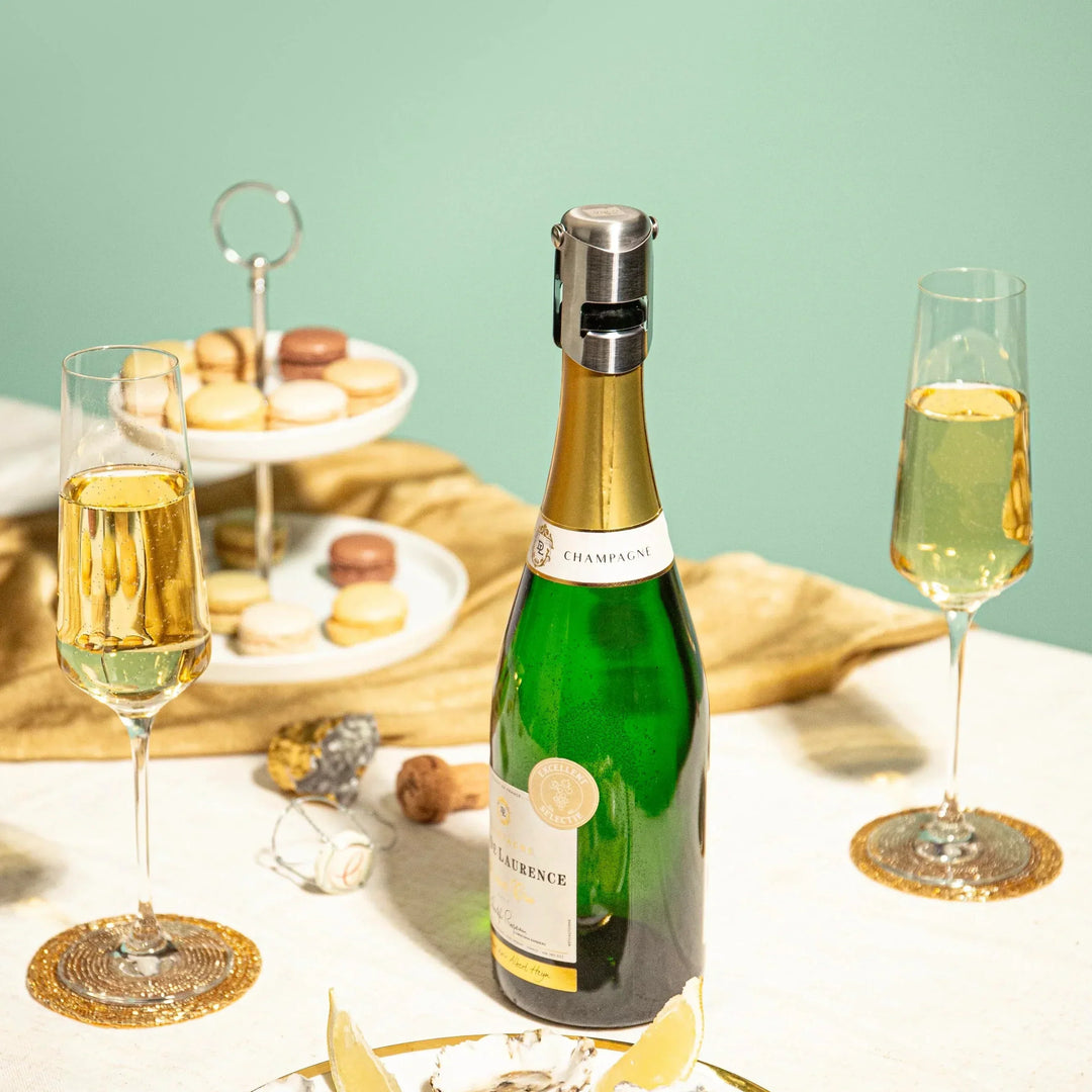Bottle of champagne with a stopper on and two glasses on a table with pastries in the background