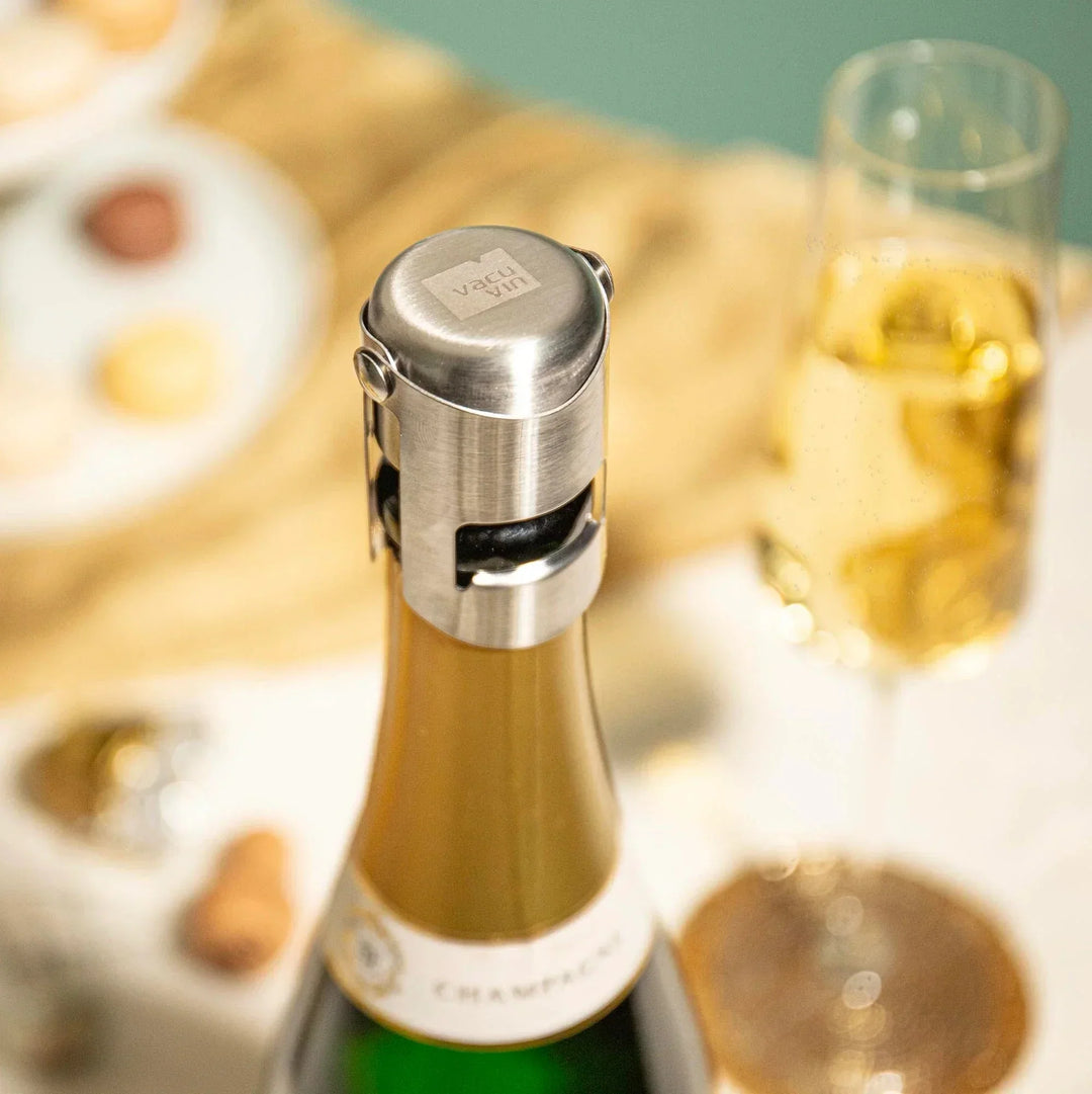 Bottle of champagne with a silver stopper on a table with a glass of champagne and snacks.
