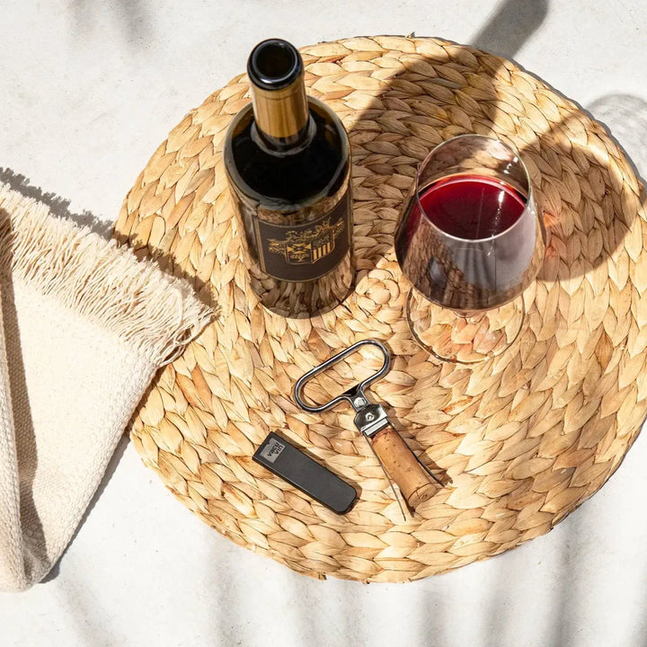Wine bottle, glass, and opener on a woven tray with a white background