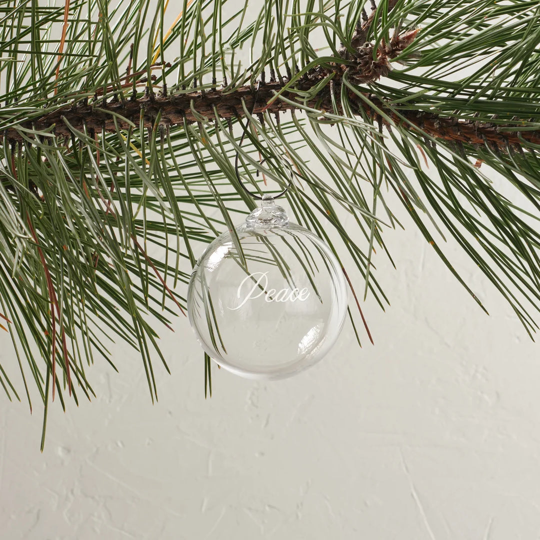 A clear glass Christmas ornament hanging from a pine tree branch, with the word 'Peace' written in elegant white script on the front.