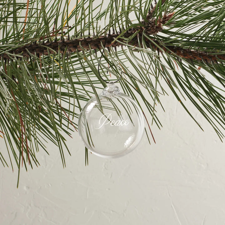 A clear glass Christmas ornament hanging from a pine tree branch, with the word 'Peace' written in elegant white script on the front.
