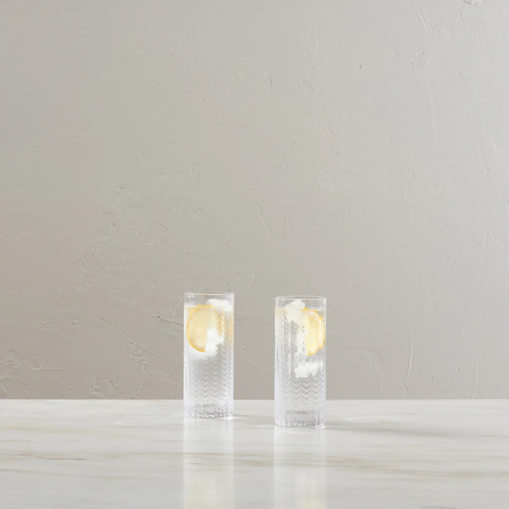 Two clear textured highball glasses filled with a clear drink and a lemon slice, set on a light marble surface against a neutral beige textured wall.
