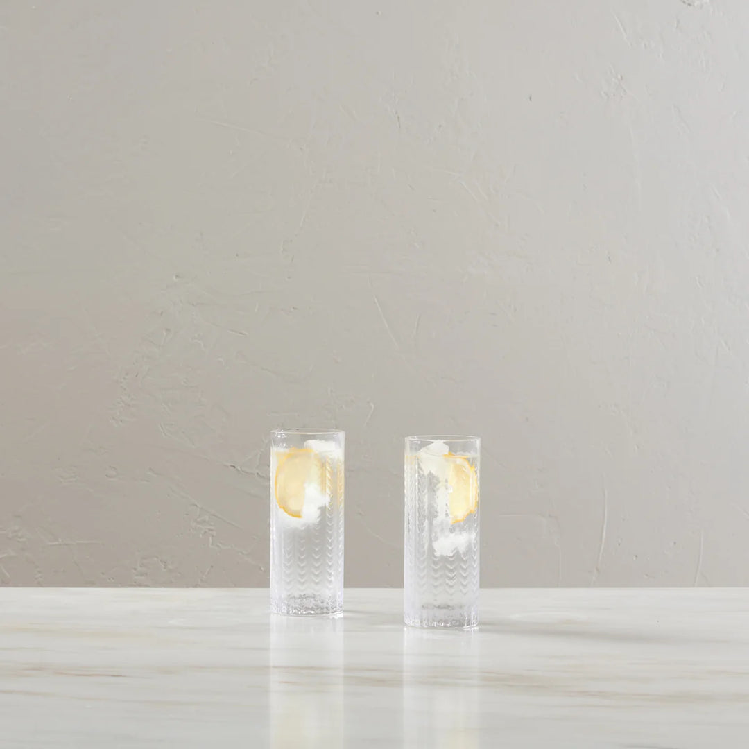 Two clear textured highball glasses filled with a clear drink and a lemon slice, set on a light marble surface against a neutral beige textured wall.