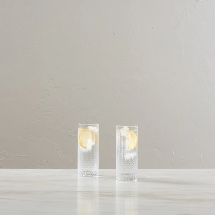 Two clear textured highball glasses filled with a clear drink and a lemon slice, set on a light marble surface against a neutral beige textured wall.